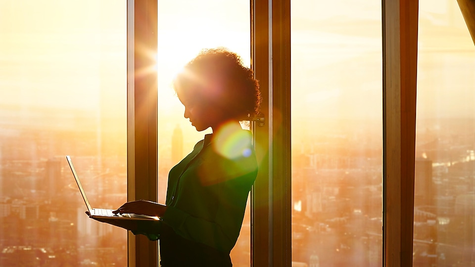 Жінка з ноутбуком стоїть у будинку перед вікном на тлі заходу сонця